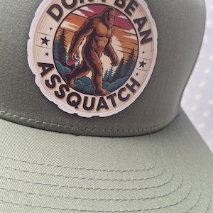 Puede incluir: Gorra de béisbol verde salvia con un parche circular con una ilustración de Bigfoot. El parche tiene el texto "DON'T BE AN ASSQUATCH" en un diseño circular con un fondo de puesta de sol y bosque.