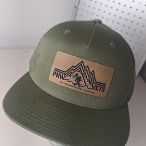 Könnte beinhalten: Eine olivgrüne Baseballkappe mit einem rechteckigen, beigefarbenen Aufnäher in Lederoptik auf der Vorderseite. Der Aufnäher zeigt eine schwarze Grafik von Bergen und einer Figur mit Flagge sowie den Text PNW VETS und PACIFIC NORTHWEST VETERANS.
