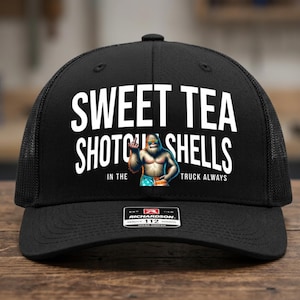 May include: A black trucker hat featuring white text that reads SWEET TEA SHOTGUN SHELLS IN THE TRUCK ALWAYS. Centered is a graphic of a hairy creature wearing sunglasses and American flag patterned shorts, holding up a middle finger gesture.