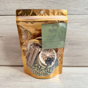May include: A gold metallic pouch filled with dried orange slices, cinnamon sticks, and chamomile. A green label reads "Rune, Earth Rose Simmer Pot, Orange, Pine, Cinnamon, Chamomile."