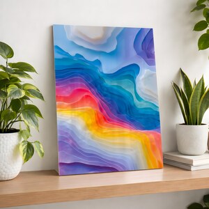 May include: A vibrant abstract art print featuring layered, colorful shapes. The artwork transitions from purple to yellow, orange, red, blue, and white, creating a gradient effect. The piece is displayed on a wooden shelf, with plants in the background.