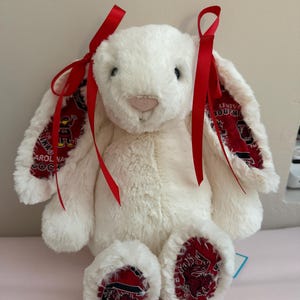 May include: A plush white bunny with red ribbon accents on its ears. The bunny's ears and footpads feature a red and black patterned fabric with the words "University of South Carolina" and a Gamecock logo. A charming stuffed animal.