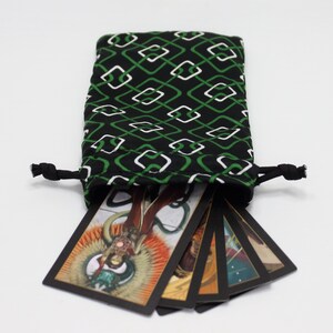 May include: A black drawstring bag with a green and white geometric pattern, holding several tarot cards. The cards have colorful illustrations and black borders. The bag is tied with black cords.