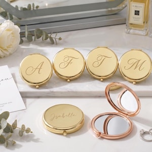 May include: A collection of round, gold-toned metal compact mirrors, some closed and engraved with letters or the name Isabella, and one open to reveal two interior mirrors. The set is displayed with white roses, a thank you card, and jewelry.