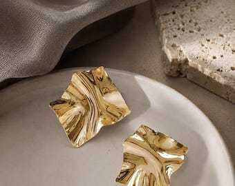 May include: A pair of gold-coloured, abstract-shaped earrings. The earrings have a textured, rippled surface and are displayed on a white plate. The background includes a neutral-toned fabric and a stone block.