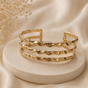 May include: A gold-toned cuff bracelet featuring three parallel bands with a wavy, hammered texture. The open-ended design consists of three distinct horizontal lines that connect at the sides, creating a layered appearance with a reflective finish.