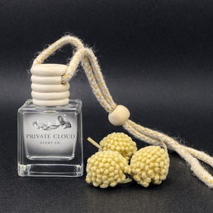 May include: A clear, square glass bottle with a wooden cap and a cream-colored rope. The bottle has a label with the text "PRIVATE CLOUD SCENT CO." and a cloud design. Three yellow, spherical decorative elements are next to the bottle.