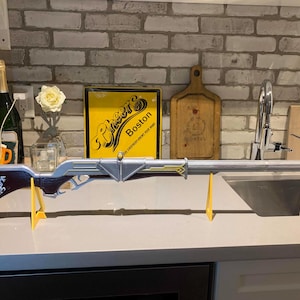May include: A silver and brown rifle prop with gold detailing, presented on a yellow stand. The prop is set against a brick wall with a "Cheers Boston" sign and a sign that says "beer is good! free beer better!".
