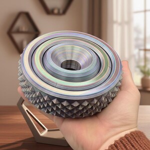 Könnte beinhalten: Ein mehrschichtiges, irisierendes Deko-Objekt mit zentralem Loch und geometrischer Basis. Das Objekt wird in einer Hand gehalten, wodurch sein kompliziertes Design und seine Farbvariationen zur Geltung kommen. Das Objekt scheint 3D-gedruckt zu sein. Der Durchmesser beträgt ca. 10 cm.