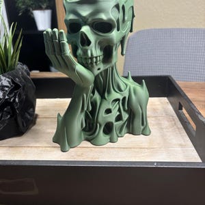 May include: A green skull-shaped decorative planter with a crown and hand supporting the chin. The planter has a detailed, textured design and is placed on a wooden tray.