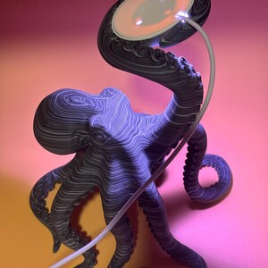 May include: A unique lamp shaped like an octopus, with a textured, layered design in shades of gray and purple. The lamp's head features a glowing, circular light with three small bulbs. A white cord extends from the lamp, set against a gradient pink and orange background.