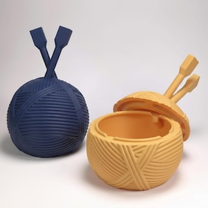 May include: Two yarn ball-shaped containers, one navy blue and one yellow, with matching knitting needle lids. The yellow container is open, revealing the interior. The containers are designed to resemble balls of yarn.