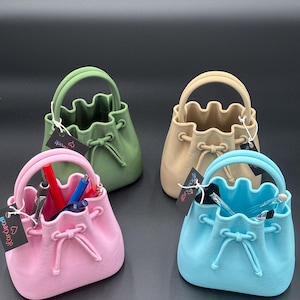 May include: Four small, purse-shaped containers in pastel colors: green, beige, pink, and blue. Each has a handle and a drawstring closure. The pink and blue purses contain pens and pencils. Each purse has a small tag attached.