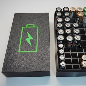 Könnte beinhalten: Ein schwarzer Batterie-Organizer mit einem Deckel mit einem grünen Batteriesymbol. Der offene Organizer zeigt Batterien verschiedener Größen. Die Batterien sind in einem Raster angeordnet und bieten eine organisierte Aufbewahrung für AA-, AAA-, C- und D-Batterien.