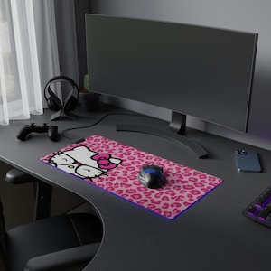 May include: A pink and white Hello Kitty mousepad with a leopard print design. The mousepad features a cartoon image of Hello Kitty wearing glasses and a bow. A computer mouse sits on the pad.