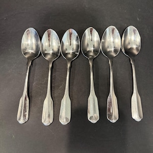 May include: A set of six silver-toned metal spoons arranged in a row. Each spoon features an oval bowl and a slender handle that widens toward the base, which is decorated with a small, fan-shaped embossed pattern at the very tip.