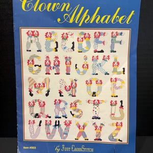 May include: A blue paper pattern sheet titled Clown Alphabet by Just CrossStitch, item #503. The sheet displays a full alphabet where each letter is formed by a colorful clown character wearing patterned clothing, including stripes and polka dots.