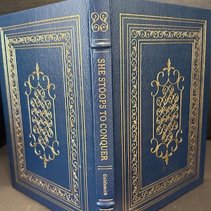 May include: A blue hardcover book titled She Stoops to Conquer by Goldsmith. The cover features gold foil embossed filigree patterns and rectangular borders. The spine displays the title and author name in gold lettering with decorative gold accents.