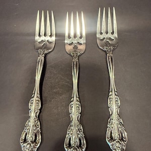 May include: Three silver-toned metal forks featuring four tines and intricate embossed floral scrollwork patterns along the handles. The design includes a decorative cutout detail at the base of the tines and a tapered handle with a rounded end.