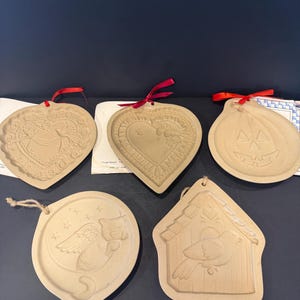 May include: A collection of beige-colored ceramic ornaments in various shapes, including a heart, pumpkin, birdhouse, and an owl. Each ornament has a detailed embossed design and is strung with a red ribbon or twine for hanging. The ornaments are likely for seasonal or decorative use.