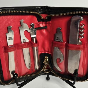 May include: A black zippered case with a bright red interior containing five stainless steel bar tools. The tools include a corkscrew, bottle openers, and various flat metal implements, each held in place by clear plastic slots inside the case.