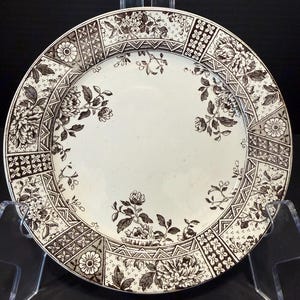 May include: A round ceramic plate featuring a cream-colored center with dark brown floral illustrations. The wide rim is decorated with a repeating pattern of geometric shapes, flowers, and leaves in a dark brown transferware style.