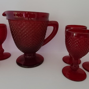 May include: A set of ruby red glassware consisting of one pitcher and five stemmed goblets. Each piece features a textured diamond pattern across the exterior surface, a circular base, and a deep, translucent red color throughout the glass.
