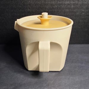 May include: A cream-colored plastic pitcher with a matching lid and a central yellow knob. The pitcher features a sturdy side handle and a small pouring spout at the top rim. The design is simple and functional, with a smooth finish and a cylindrical body shape that tapers slightly toward the base.