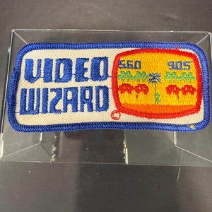 May include: A rectangular embroidered patch with a blue border on a beige background. The text "VIDEO WIZARD" is in blue. To the right is a red-outlined yellow screen graphic featuring green pixelated trees, red pixelated shapes, and blue numbers.