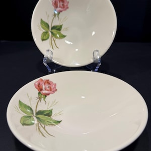 May include: Two off-white ceramic bowls with a pink rose and green leaves design. The bowls are round and have a shallow depth. The rose is in full bloom, and the leaves are detailed with green and brown shading.