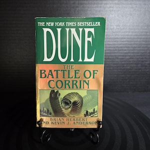 May include: A paperback book titled Dune: The Battle of Corrin by Brian Herbert and Kevin J. Anderson. The cover features a green top section with white text, a gold middle section, and an illustration of a large sandworm and figures.