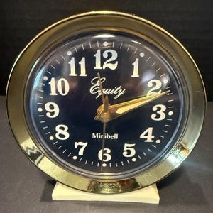 May include: A round analog alarm clock with a polished gold-tone metal frame and a dark blue face. The clock features white Arabic numerals, gold-tone hands, and the text Equity and Minibell printed in white on the face. It sits on a white base.