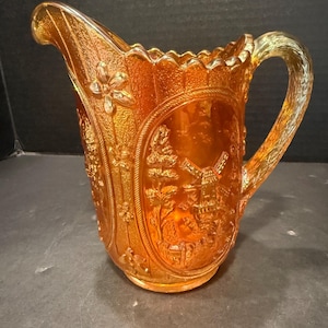 May include: An orange iridescent carnival glass pitcher featuring a relief pattern of a windmill in a landscape scene, surrounded by floral motifs. The pitcher has a scalloped rim, a textured handle, and a glossy finish that reflects light.