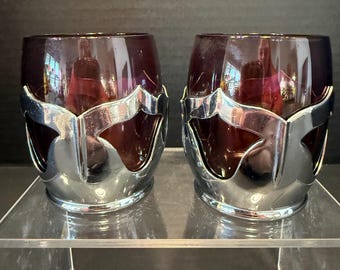 May include: Two deep purple glass tumblers, each encased in a polished silver-tone metal holder featuring a decorative cutout design. The holders wrap around the lower portion of the rounded glass vessels, creating a contrast between the dark glass and the reflective metallic finish.