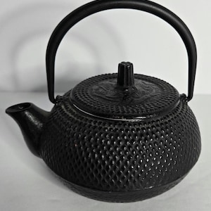 May include: A black cast iron teapot featuring a textured hobnail pattern across its rounded body. The teapot includes a matching lid with a small central knob and a curved handle that arches over the top. A short spout extends from the side.