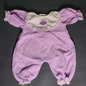 May include: A lavender baby onesie with a white collar and lace trim. The collar features a purple embroidered floral design. The sleeves and legs have ruffled lace edges. The onesie is made of a soft, plush material.