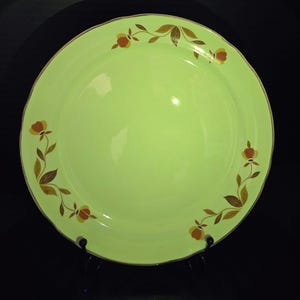 May include: A round, light green ceramic dinner plate featuring a scalloped edge and a decorative border of brown and orange floral motifs. The plate has a smooth, glossy finish and is displayed on a black stand against a dark background.