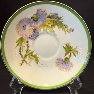 May include: A white porcelain saucer featuring a hand-painted design of purple thistle flowers with green leaves and stems. The rim is accented with a bright green band and a thin gold edge. The signature P. Curnock is visible near the floral motif.