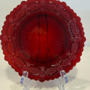 May include: A round, deep red glass bowl featuring a scalloped edge and a decorative embossed pattern along the rim. The interior of the bowl displays a radial sunburst design that extends from the center toward the textured, patterned border.