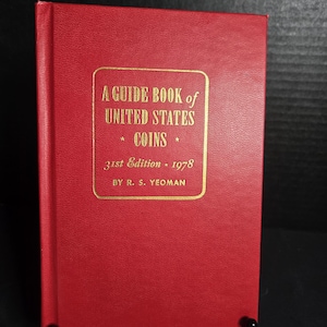 May include: A red hardcover book titled A GUIDE BOOK of UNITED STATES COINS 31st Edition 1978 BY R. S. YEOMAN. The front cover features gold embossed text centered within a gold rectangular border, with a textured finish on the red cover material.