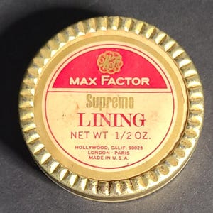 May include: A round, gold-colored metal container lid with a scalloped edge. The label features a red and cream background with the text MAX FACTOR, Supreme LINING, NET WT 1/2 OZ., HOLLYWOOD, CALIF. 90028, LONDON, PARIS, and MADE IN U.S.A.