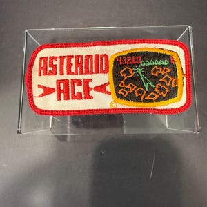 May include: A rectangular embroidered patch with a red border on a white background. The text ASTEROID ACE is in red. To the right is a black screen graphic with orange asteroid shapes, a green radar icon, and the red numbers 43210.