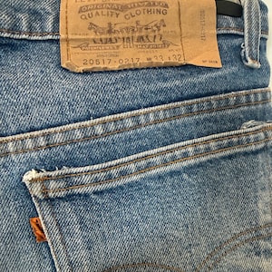 May include: Blue denim jeans featuring a tan leather patch on the waistband with the text Levi Strauss & Co. Original Riveted Quality Clothing 20517-0217 W 33 L 32. The jeans include a back pocket with an orange tab and brown contrast stitching.