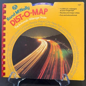 May include: A vintage Rand McNally DIST-O-MAP, an automatic mileage dialer, with a bright yellow cover and red spiral binding. The cover features a highway image with light trails and text including "11,000 U.S. mileages at your fingertips."