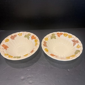 May include: Two cream-colored ceramic bowls with a scalloped rim and a fluted interior. The exterior rim features a repeating pattern of small yellow flowers, green leaves, and brown autumn leaves painted around the circumference of each bowl.