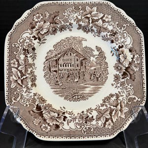 May include: A square ceramic plate with rounded corners featuring a brown and white transferware pattern. The center depicts a house and figures in a landscape, surrounded by a wide border of detailed floral motifs and geometric lattice.