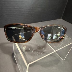 May include: Tortoiseshell-patterned sunglasses with dark, polarized lenses. The left lens features the text 'Zuma 1777508038 CE' and the right lens displays a small logo with the word 'POLARIZED' printed in white lettering on the upper corner.