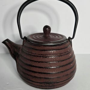 May include: A round, dark brown cast iron teapot featuring a textured surface with horizontal black stripes. The teapot includes a matching lid with a small knob handle and a tall, arched black metal handle attached to the sides of the body.