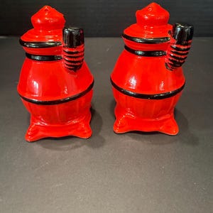 May include: A pair of bright red ceramic salt and pepper shakers shaped like vintage potbelly stoves. Each shaker features a glossy finish with black painted accents on the lid, the stovepipe, and the base, standing on four small feet.
