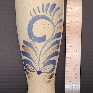 May include: A tall, flared ceramic vase with a cream-colored speckled finish, featuring blue and brown painted leaf-like motifs. A metal ruler placed alongside shows the vase stands approximately 11 inches tall, with a base diameter of 3 inches.